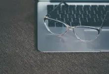 نظارة طبية شفافة موضوعة على لابتوب فضي فوق قماش رمادي
