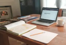 جهاز محمول بمستند مفتوح على مكتب خشبي بجانب دفاتر وكوب