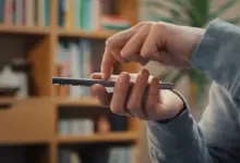 يد تمسك هاتفًا ذكيًا أمام رف كتب