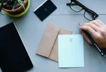 يد تكتب على ورقة بجانب مظروف ونظارة ودفتر على مكتب رمادي