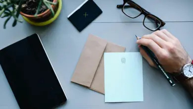 يد تكتب على ورقة بجانب مظروف ونظارة ودفتر على مكتب رمادي