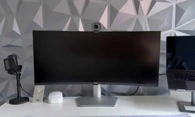 شاشة Dell منحنية عريضة على مكتب أبيض بجانب ميكروفون وكاميرا ويب