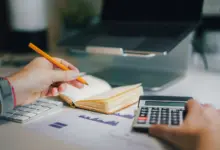 دفتر وآلة حاسبة ومخططات على مكتب أمام جهاز محمول