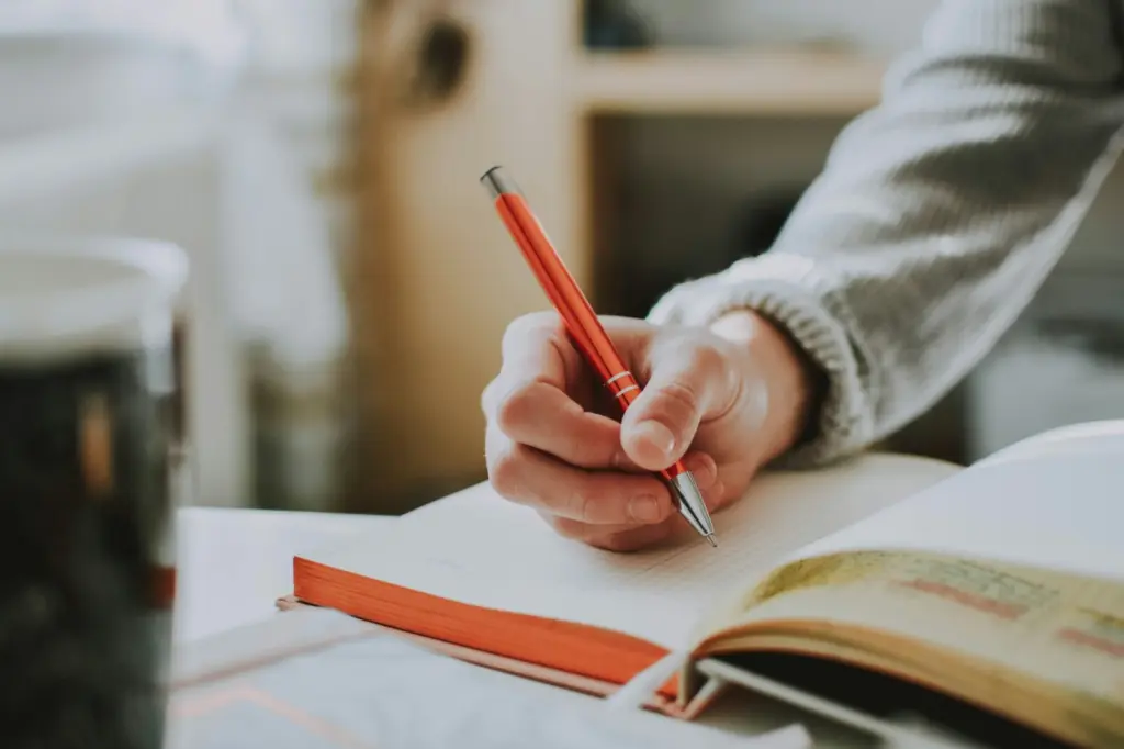 يد تكتب بقلم أحمر بجانب كتاب مفتوح على طاولة