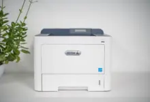 طابعة ليزر Xerox بيضاء على رف أبيض بجانب نبتة